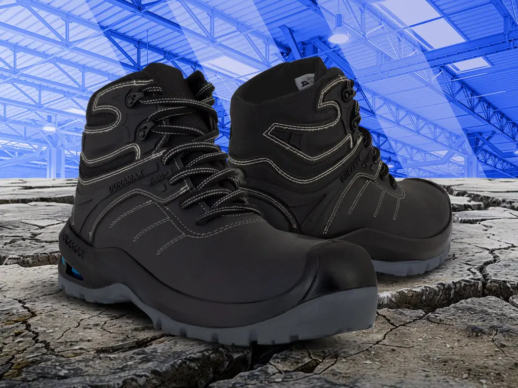Par de botas de seguridad Duramax negras sobre tierra agrietada, frente a una nave industrial