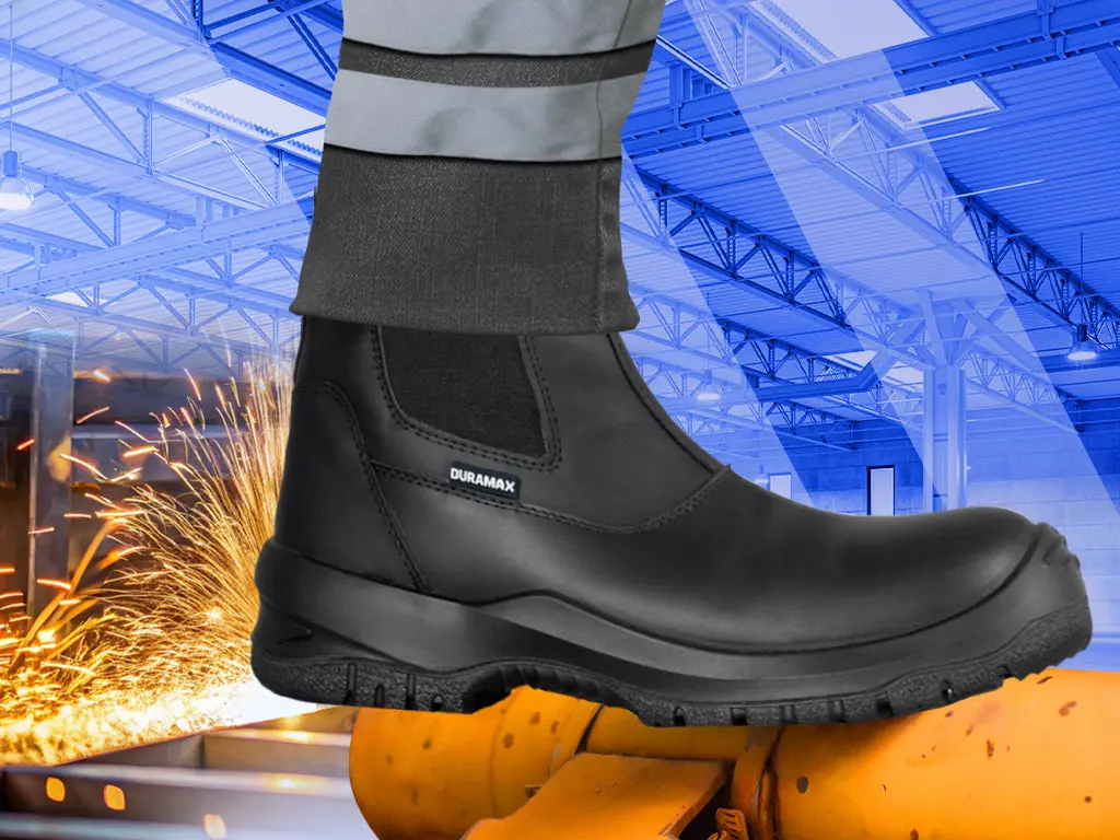 Bota de seguridad Duramax con pantalón reflejante sobre una estructura amarilla, con chispas de metalurgia al fondo